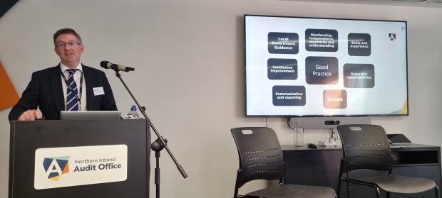NI Audit Office Director presenting to the audience on the key themes of the Effective Audit and Risk Assurance Committee guide. A presentation slide is shown listing the main sections - Good Practice, Local Government Guidance, Membership independence, objectivity and understanding Skills and experience Continuous Improvement Communication and reporting Scope Roles and responsibilities
